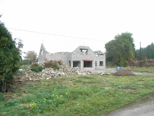 An unfinished two-storey blockwork house with open window spaces, timber roof framing and rubble in the foreground.